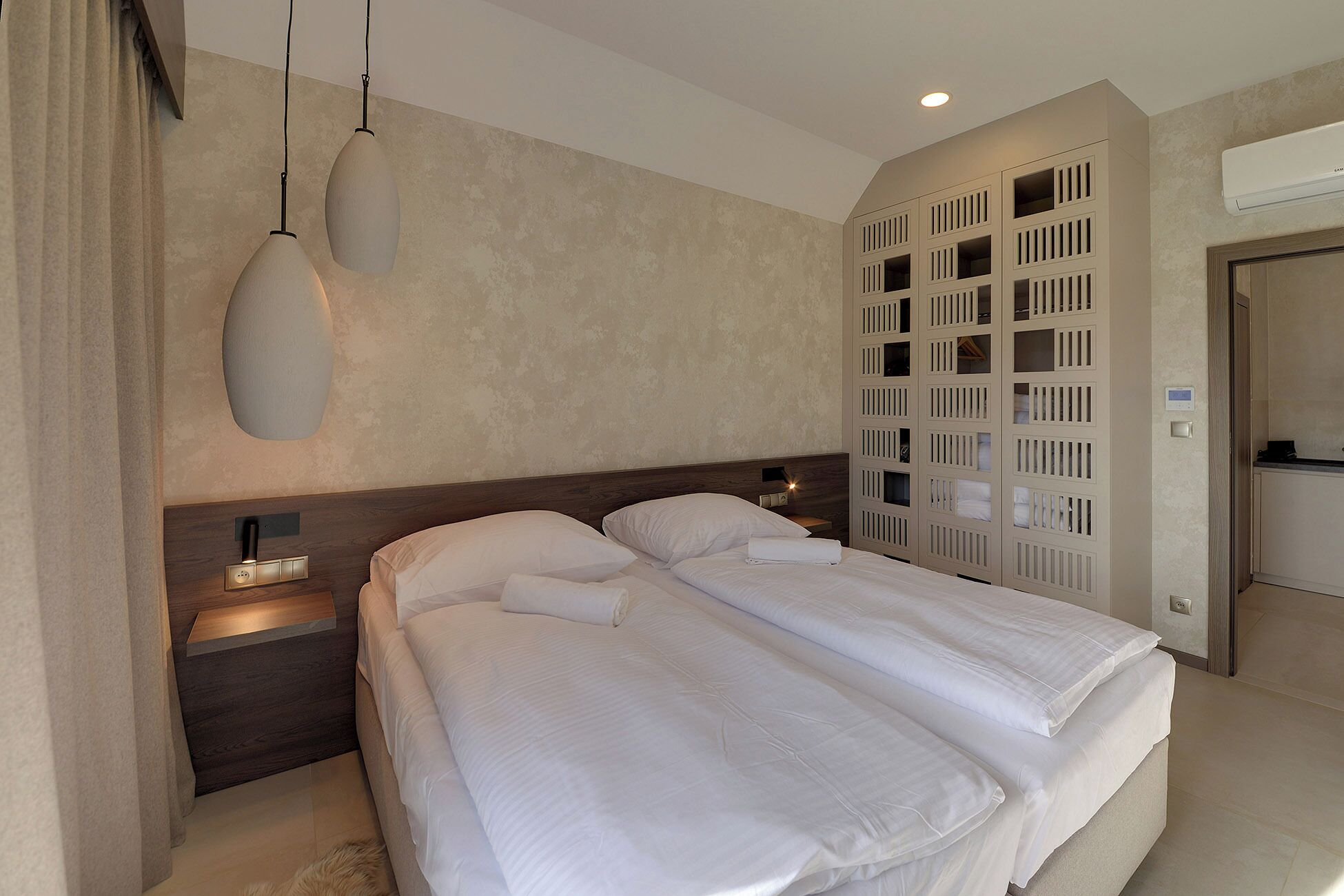 Ein Hotelzimmer mit einem großen Bett, Nachttischen, einer Hängelampe und einer eingebauten Garderobe mit mehreren Fächern.