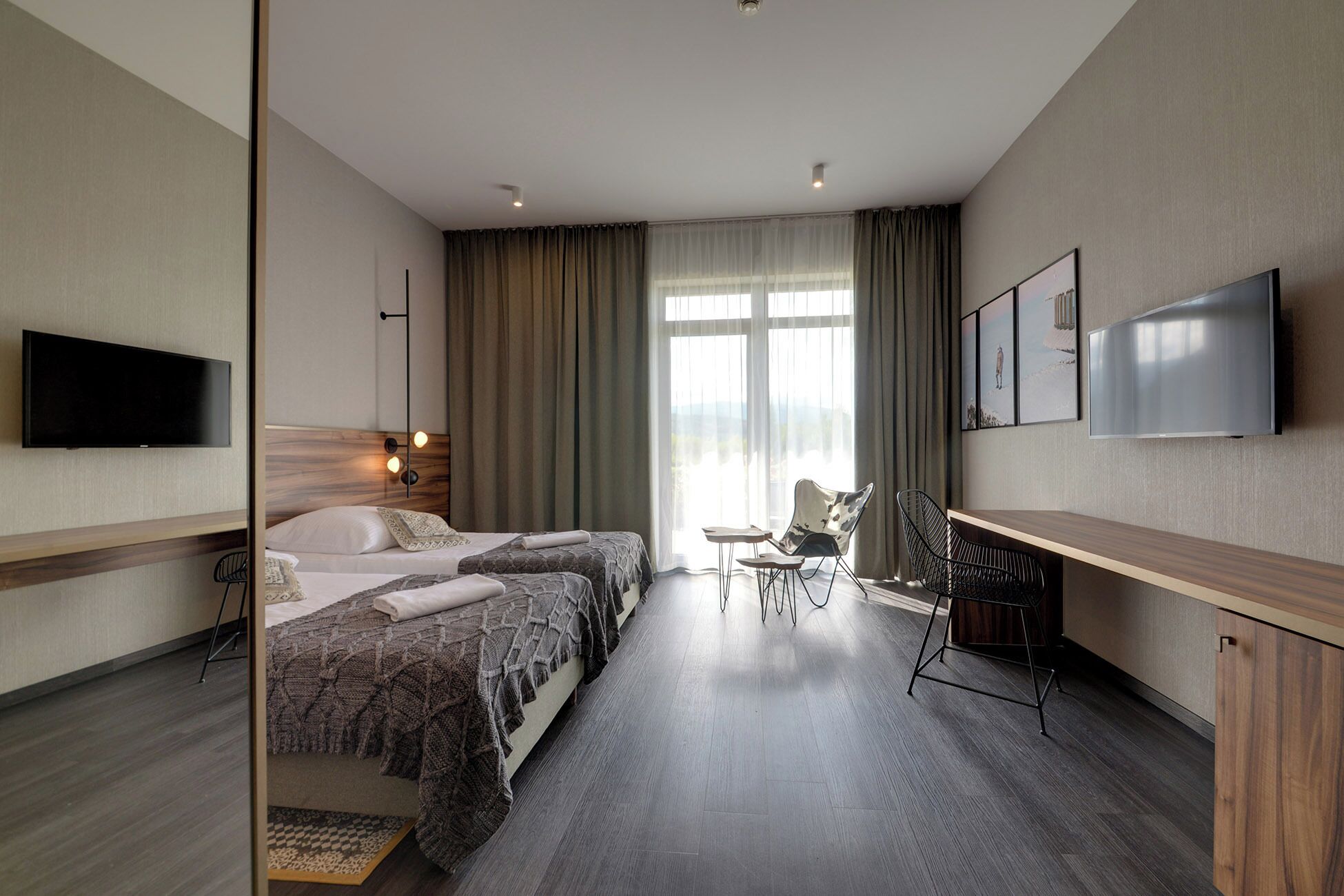 A hotel room with two beds, a desk, a chair, a TV, and a large window with curtains.