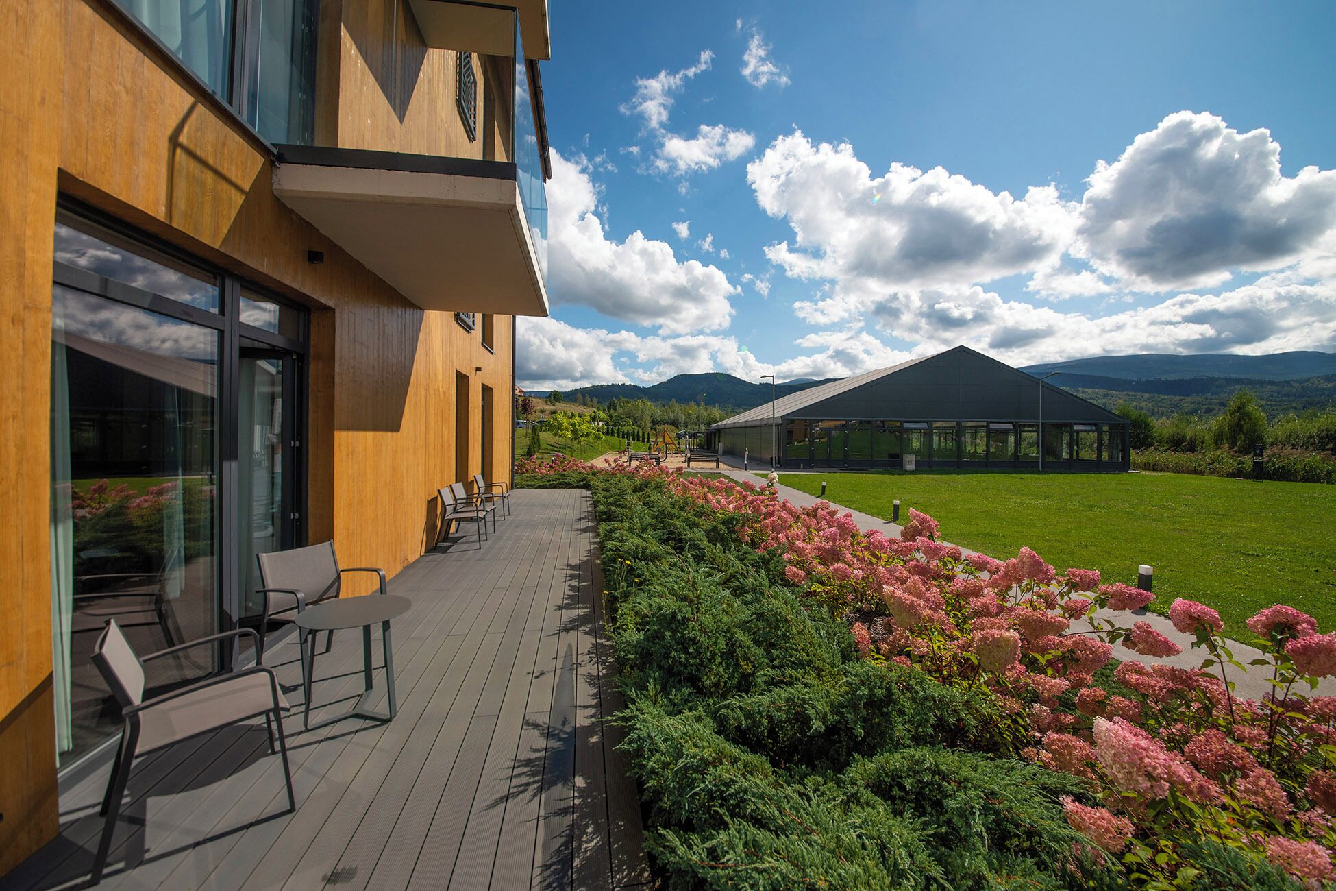 Ein modernes Gebäude mit Holzfassade und großen Fenstern, eine Terrasse mit Außenbestuhlung und Blick auf eine grüne Landschaft mit Bergen im Hintergrund.