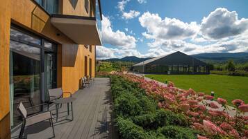 Ein modernes Gebäude mit Holzfassade und großen Fenstern, eine Terrasse mit Außenbestuhlung und Blick auf eine grüne Landschaft mit Bergen im Hintergrund.