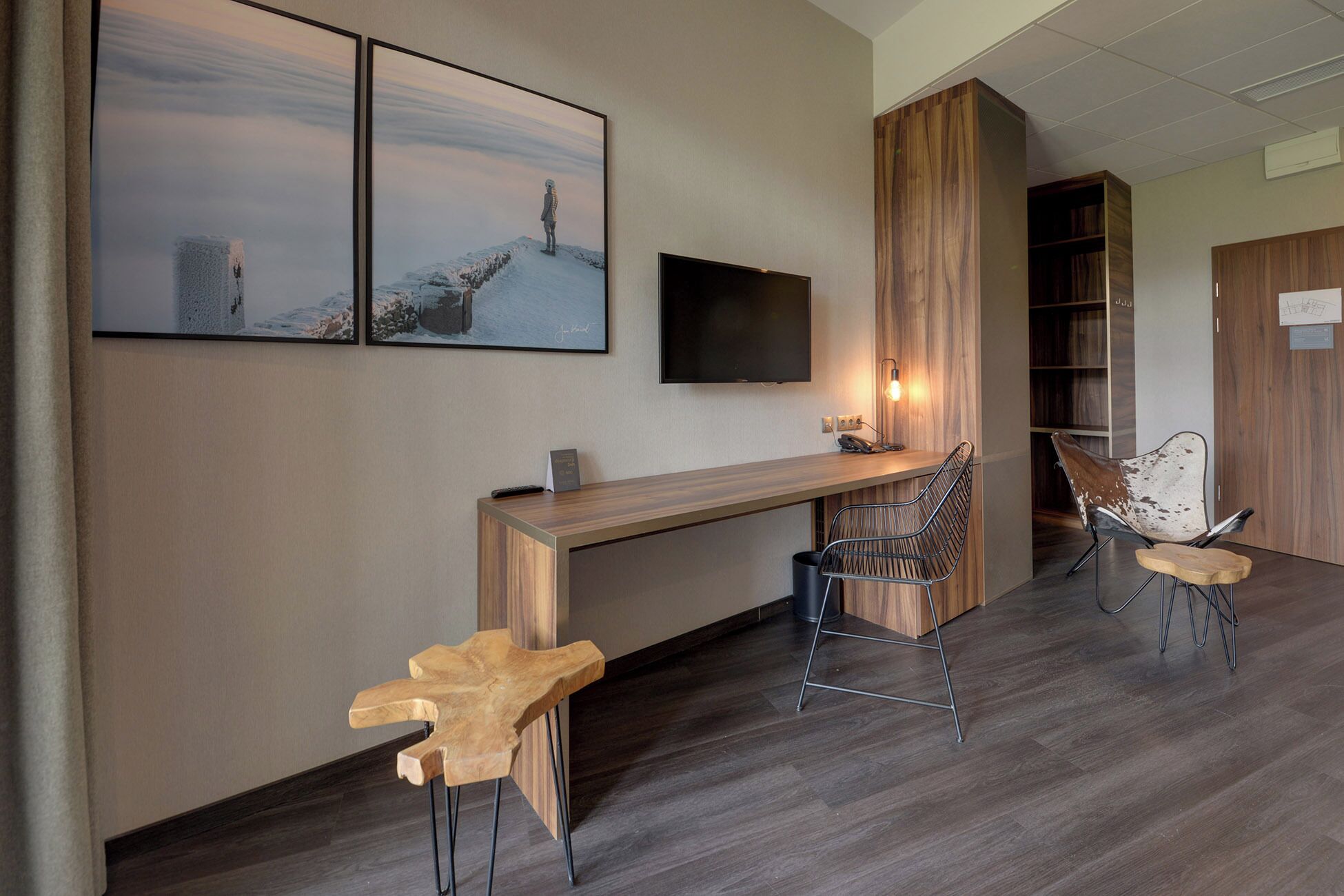 A modern hotel room with a wooden desk, two bar stools, a TV mounted on the wall, and a large window.