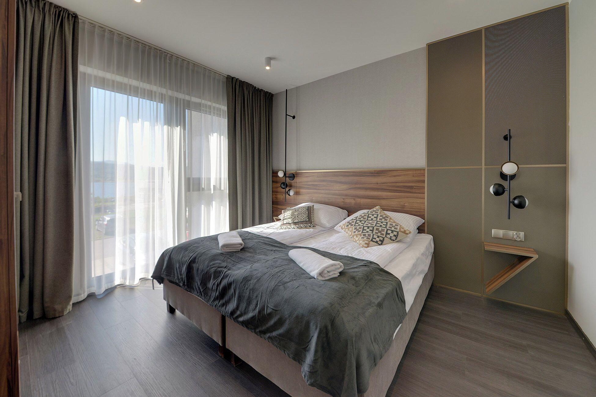 Ein modernes Hotelzimmer mit einem großen Bett, einem Holzkopfstück und Blick ins Freie durch transparente Vorhänge.