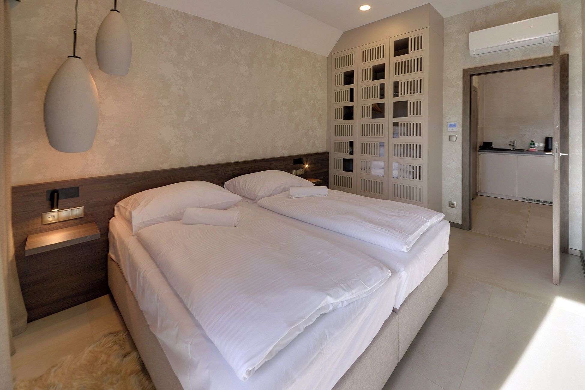 A modern hotel room with a large bed, bedside lamps, a built-in wardrobe, and a kitchenette.