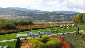 Área de estar ao ar livre à beira do lago, com mesas e cadeiras, rodeada por vegetação exuberante e vista para o lago e montanhas.