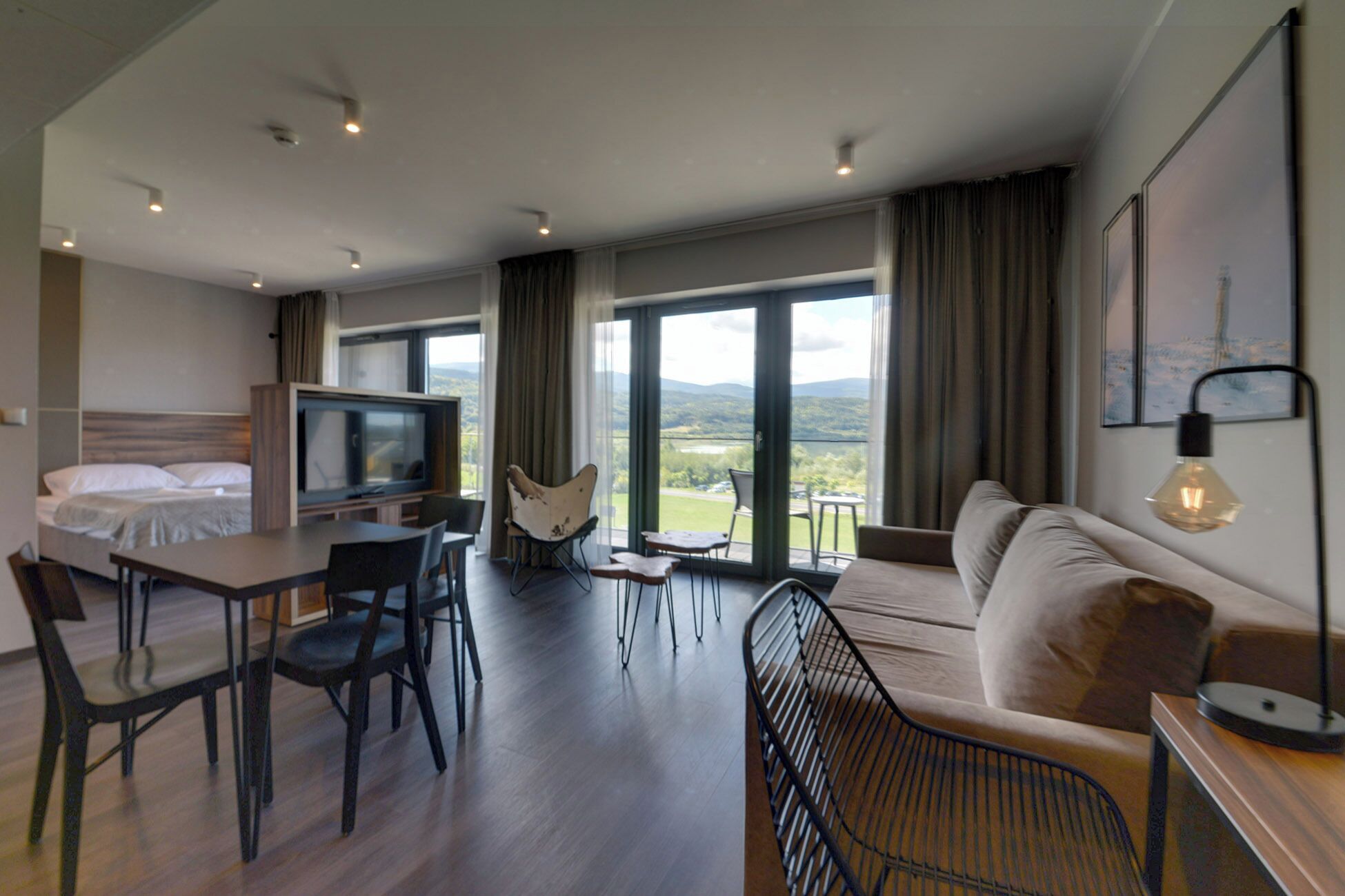 Een moderne hotelkamer met een bed, een televisie, een zithoek met stoelen, een bank en grote ramen met uitzicht.