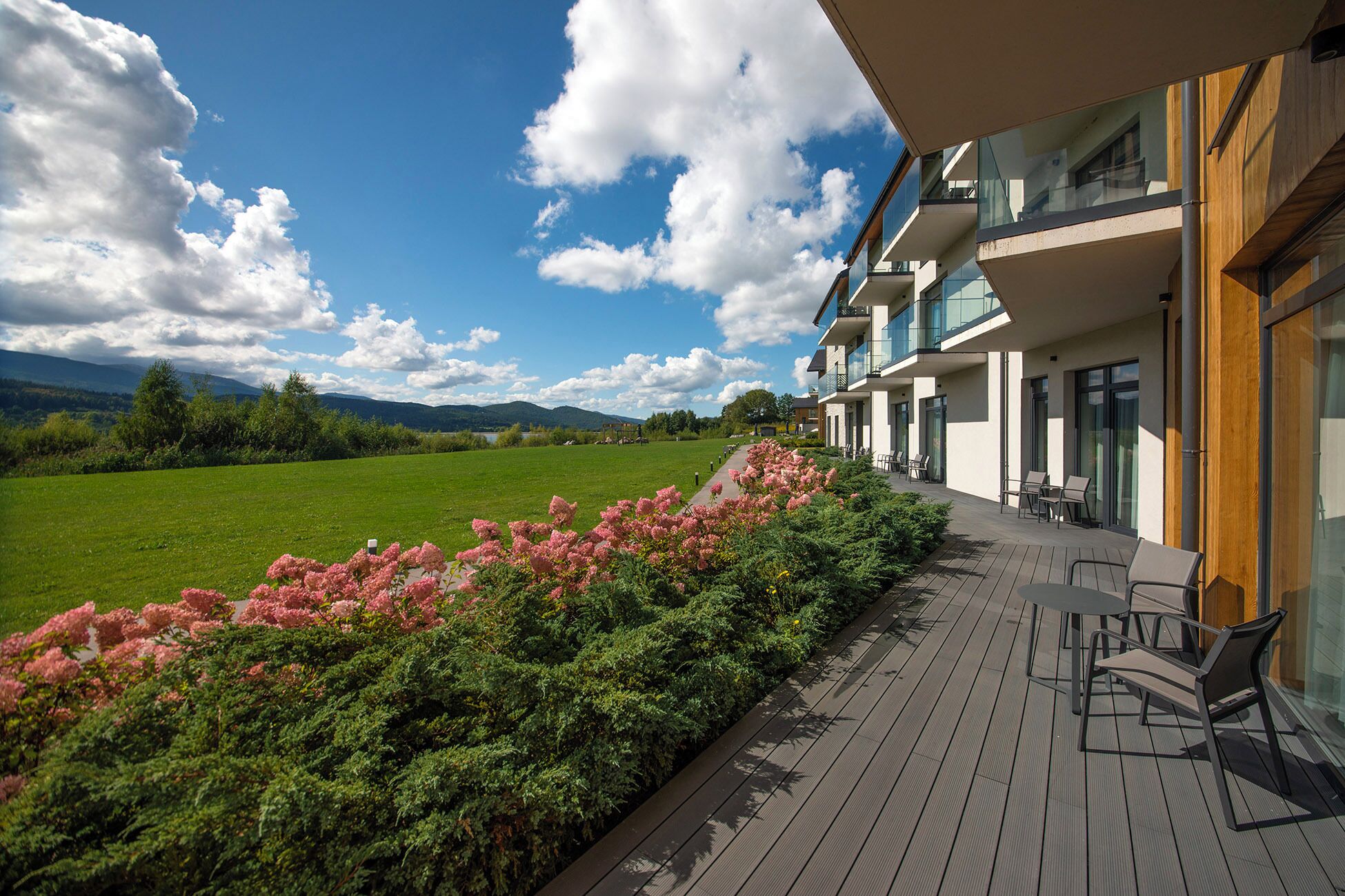 A hotel with a wooden deck, outdoor seating, and a view of a green field and mountains.