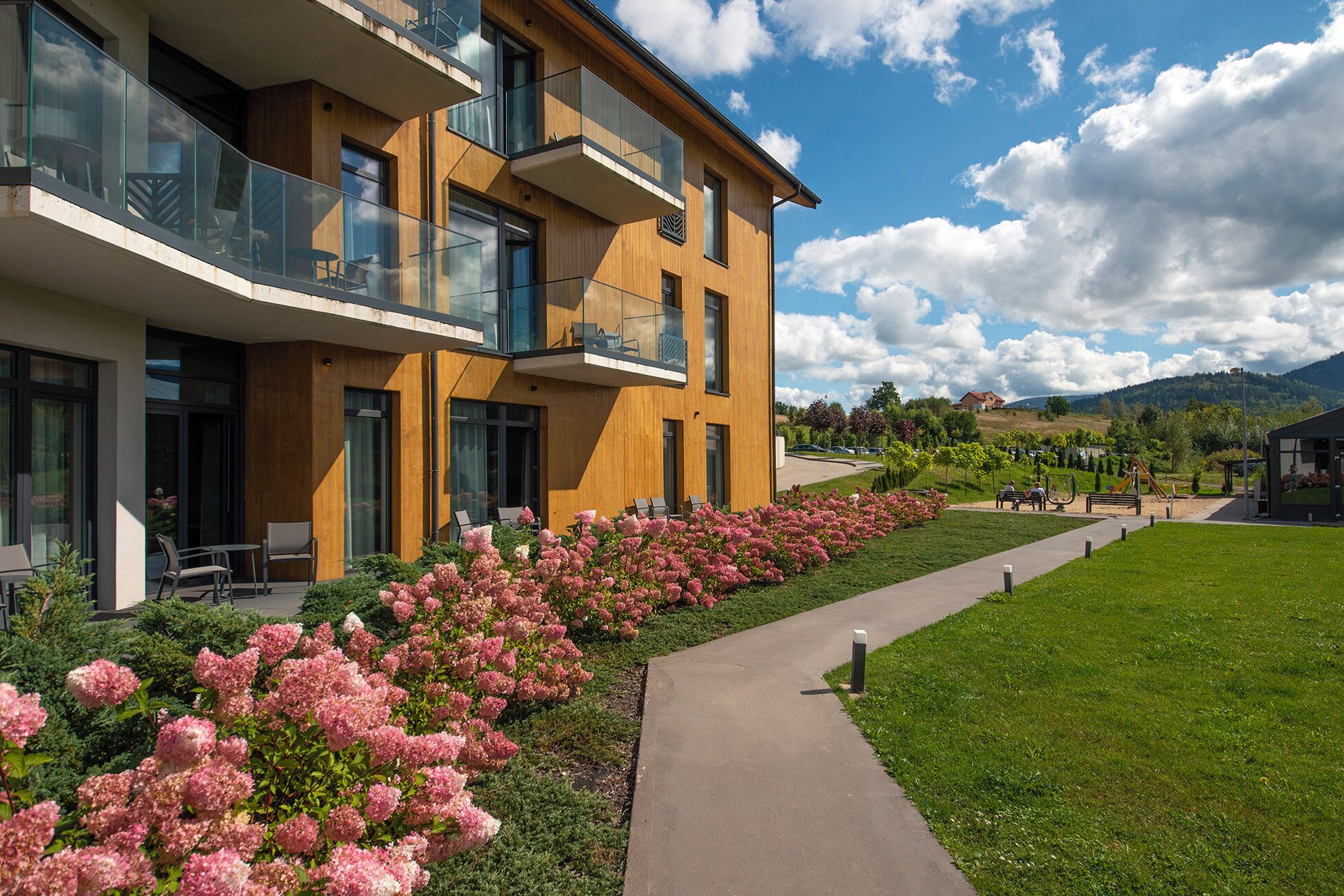 Un bâtiment moderne doté de balcons, d’une allée pavée et d’un jardin aménagé avec des fleurs roses.