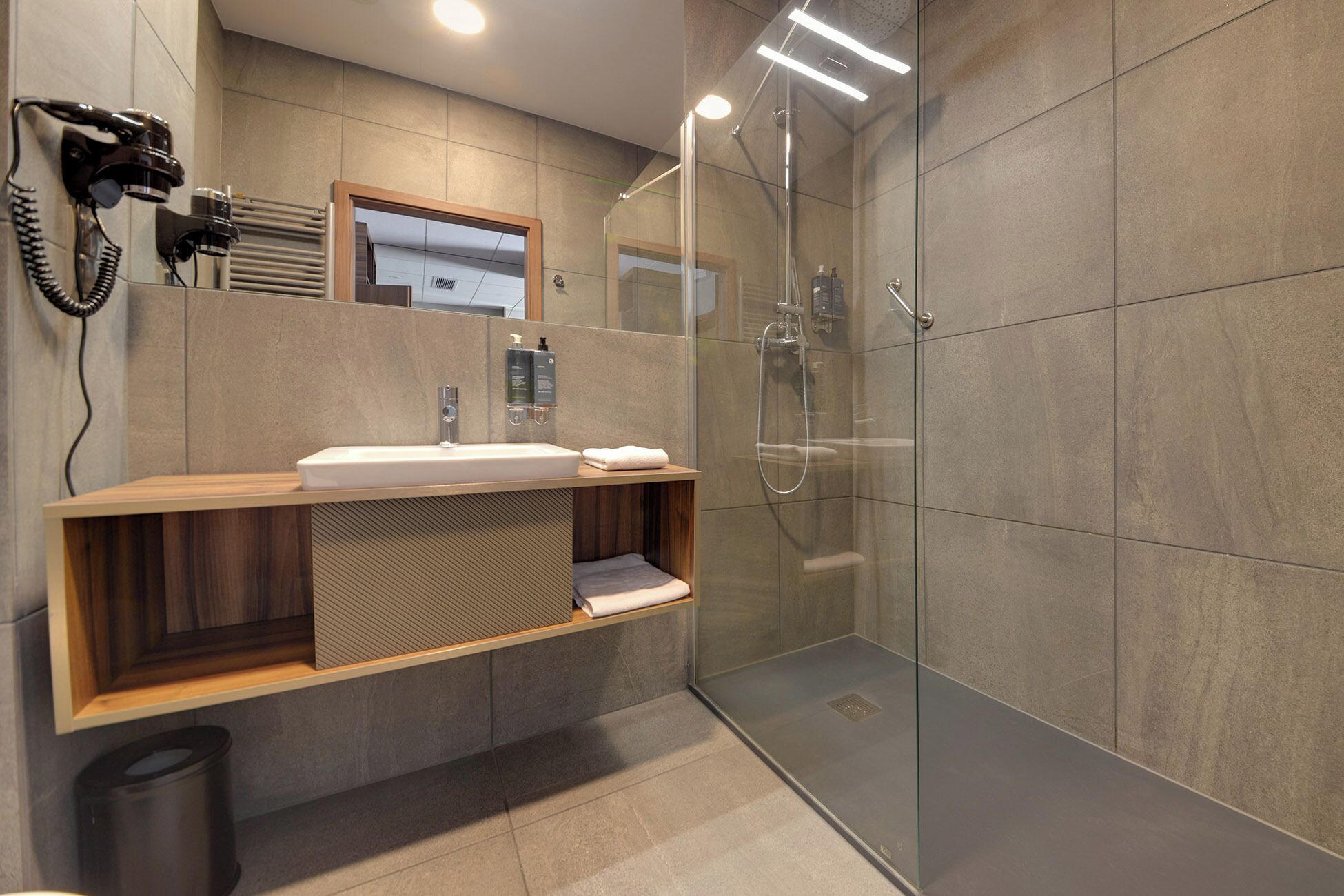 Un bagno moderno con doccia vetrata, mobile contenitore in legno con lavabo e asciugacapelli fissato alla parete.
