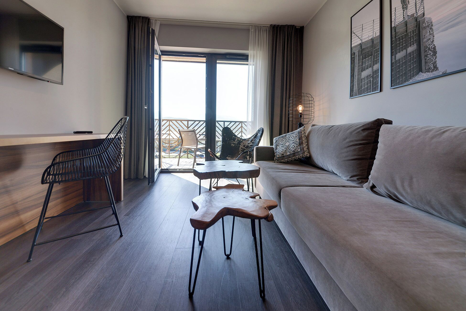 A modern hotel room with a sofa, a small table, a chair, and a balcony with outdoor seating.