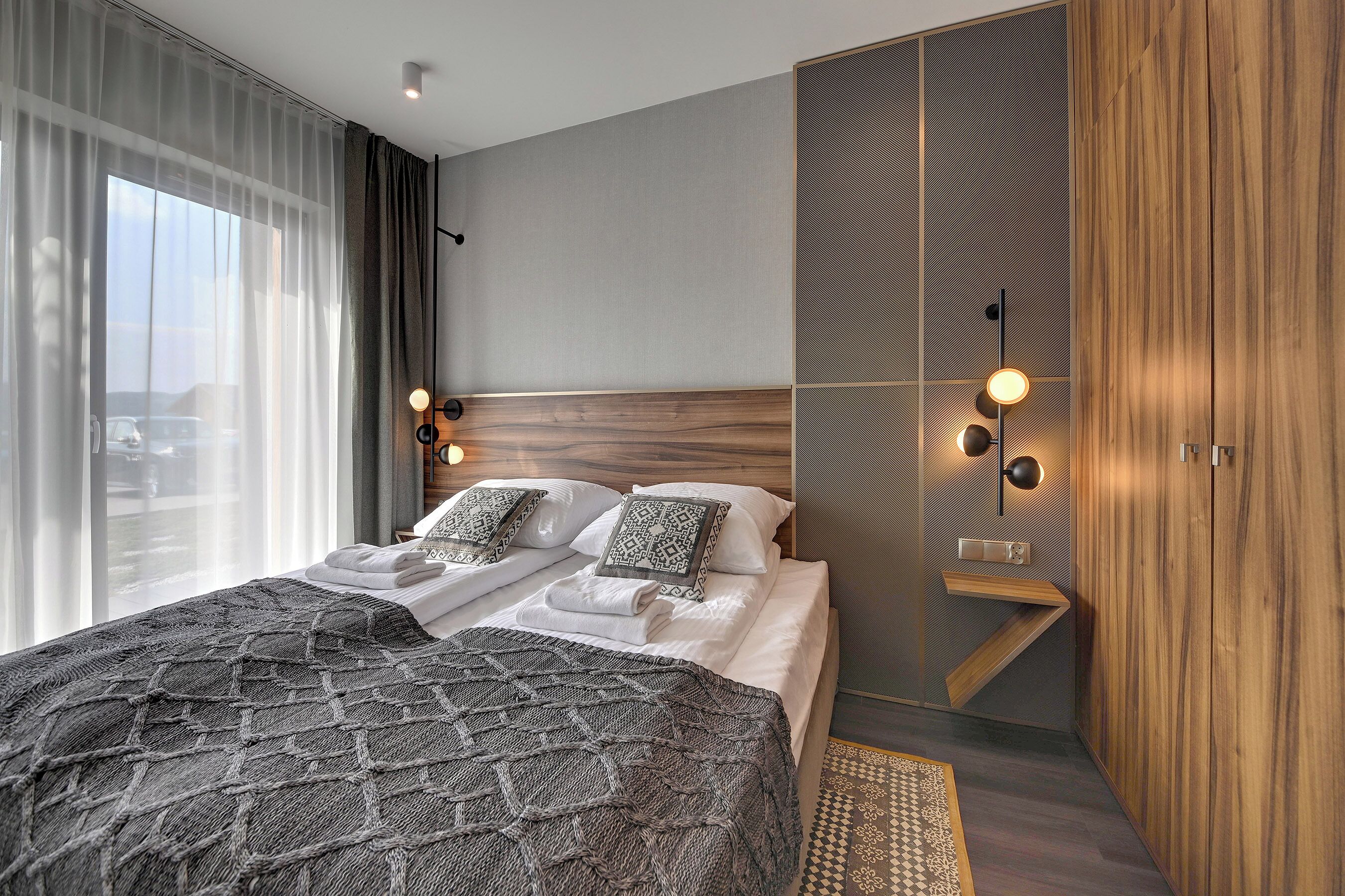 A modern hotel room with a large bed, a wooden headboard, a small seating area, and a view of the outdoors.