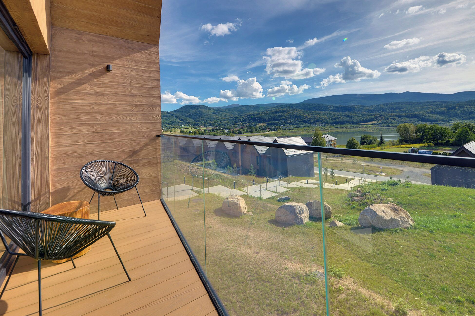 A balcony with a wooden floor, a chair, and a glass railing overlooking a scenic landscape with mountains, a lake, and buildings.