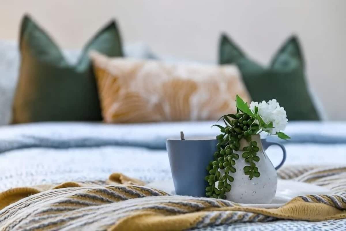 Una cama con una manta azul, un jarrón blanco con follaje verde y una taza azul.