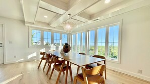 A spacious dining room with a long wooden table and chairs, large windows, and a chandelier.
