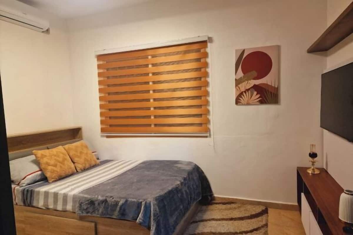 A bedroom with a bed, wooden blinds, a painting, and a TV on a stand.