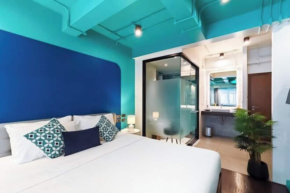 A modern hotel room with a bed, a nightstand, a lamp, a potted plant, and a bathroom visible through a glass door.