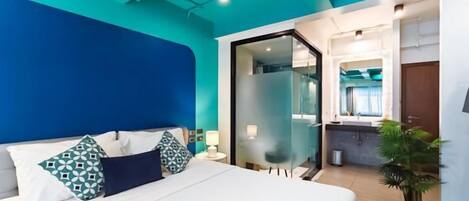 A modern hotel room with a large bed, a glass door leading to a bathroom, and a potted plant.