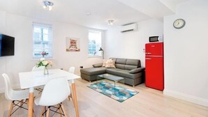 Un salon moderne avec un canapé d’angle, une table à manger avec des chaises, un réfrigérateur rouge et une horloge au mur.