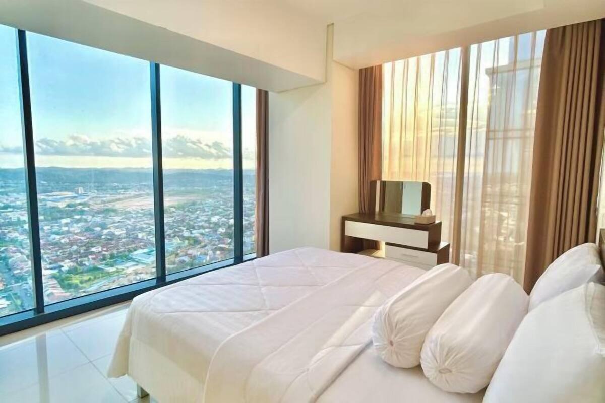 A bedroom with a large window, a bed, a bedside table, and a view of a cityscape.
