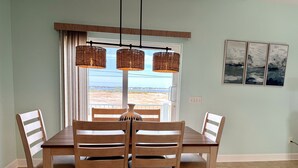 A dining area with a wooden table and chairs, a view of the beach through a large window, and a hanging light fixture. - Seaside Bungalow 1 Bedroom Home (Navarre)