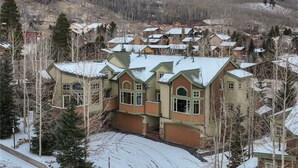 A large, multi-story house with a prominent front porch and a garage, surrounded by snow-covered trees and a hilly backdrop. - 24 Autumn Brook 4 Bedroom Townhouse (Keystone)