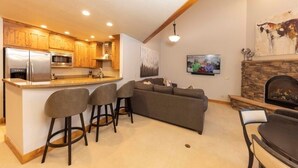 A modern kitchen with wooden cabinets, a built-in island with bar stools, a gray sofa, and a stone fireplace. - 24 Autumn Brook 4 Bedroom Townhouse (Keystone)