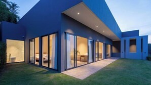 A modern house with large glass doors leading to a well-lit interior, a neatly trimmed lawn, and a clear sky. - THE GREEN LOTUS SAMUI (Koh Samui)