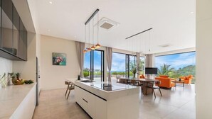 A modern kitchen with a central island, dining area with orange chairs, and large windows offering a view of greenery. - THE GREEN LOTUS SAMUI (Koh Samui)