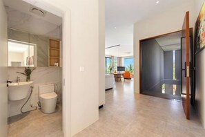 Un baño moderno con lavamanos y retrete de mármol, un pasillo que conduce a una sala de estar con un ventanal grande, y una puerta con vistas al exterior.