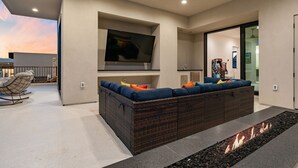A modern living room with a large flat-screen TV, a wicker sofa with colorful cushions, a fire feature, and a glass door leading to another room.
