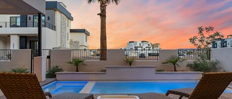 A poolside area with lounge chairs and a palm tree, overlooking a modern building complex.