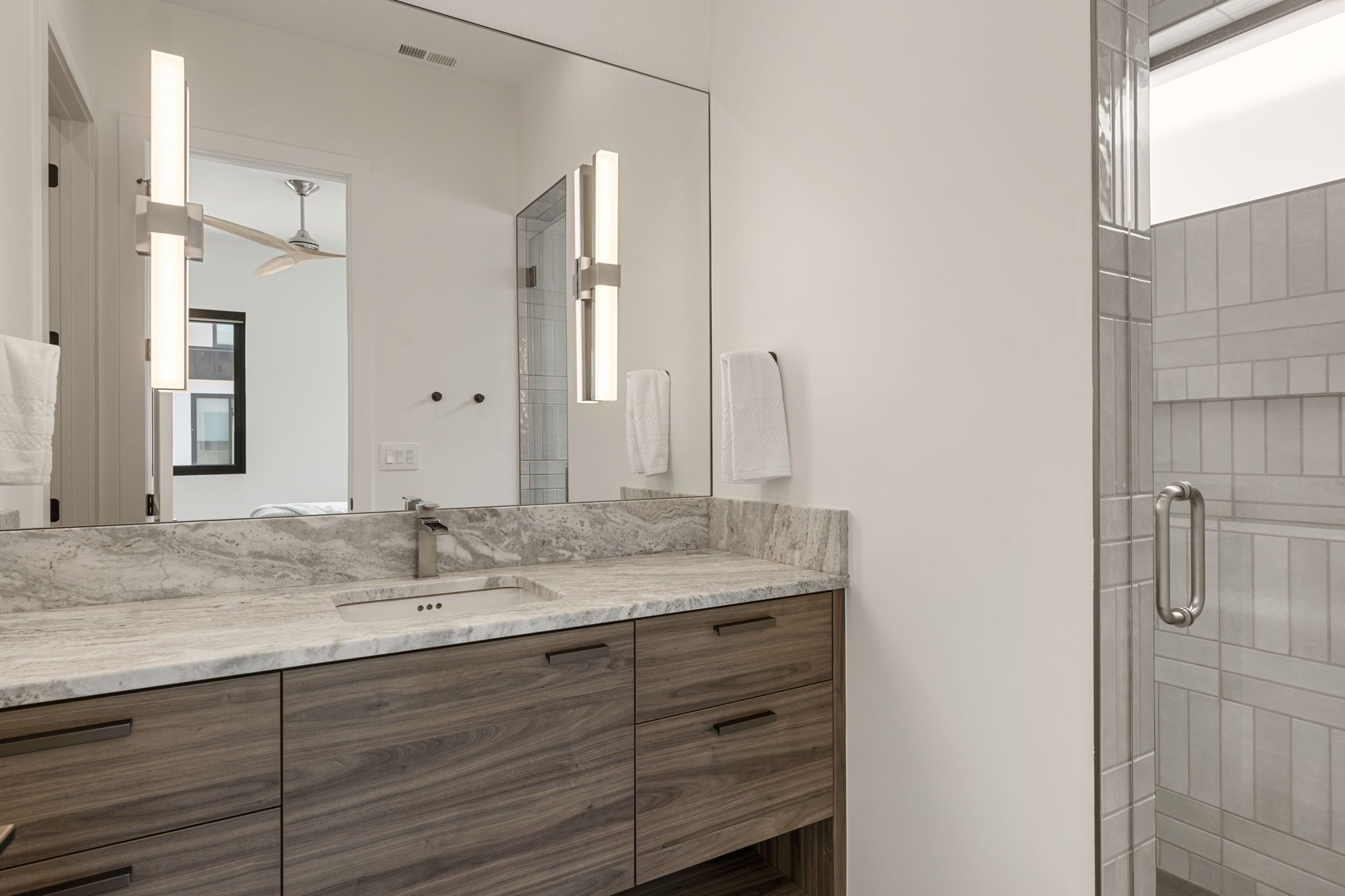 A modern bathroom with a marble countertop, wooden cabinets, a large mirror, and a glass shower enclosure.