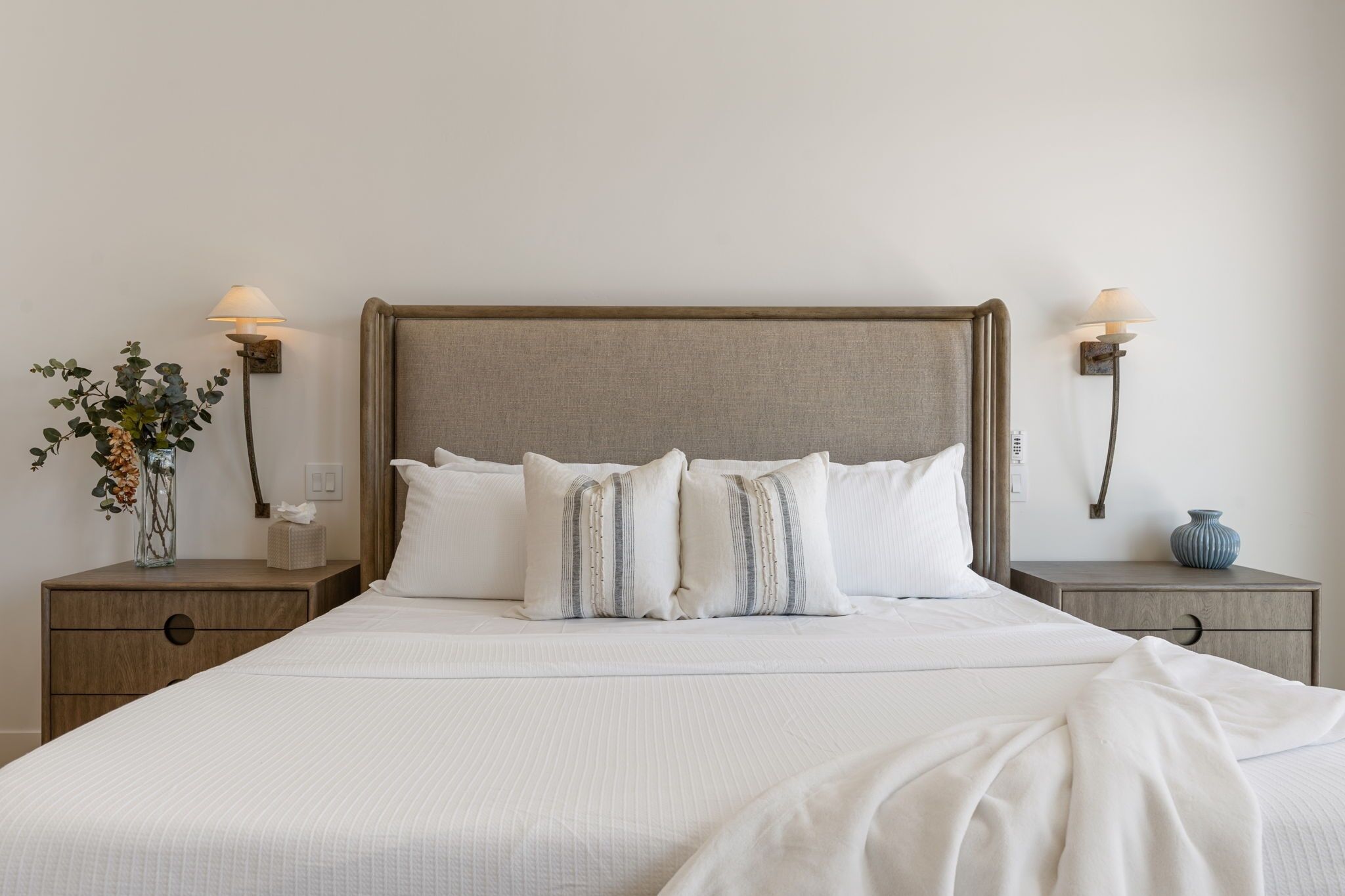 Una cama bien tendida con un cabecero de madera, dos mesitas de noche con lámparas, y un jarrón con flores en una de ellas.