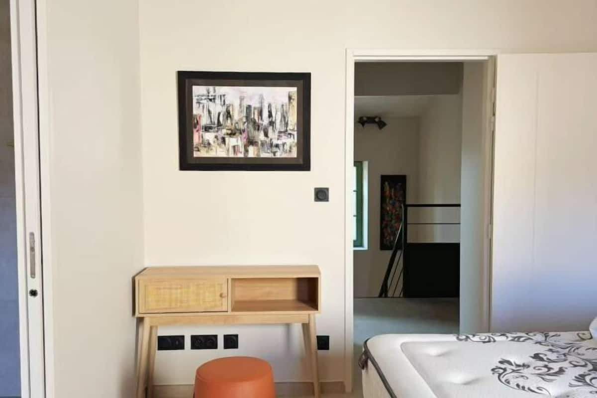 Ein Schlafzimmer mit einem Bett, einem kleinen Holztisch, einem orangefarbenen Hocker und einem gerahmten abstrakten Gemälde an der Wand.