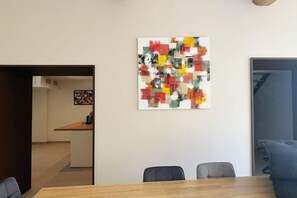 Un coin repas avec une table et des chaises en bois, un tableau abstrait aux couleurs vives accroché au mur et une vue sur la cuisine.