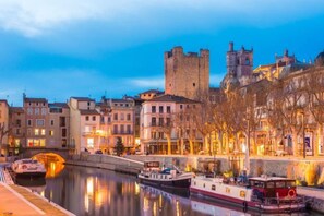 Une vue urbaine le long d’une rivière, avec des bâtiments historiques, un château et un bateau amarré au bord de l’eau.