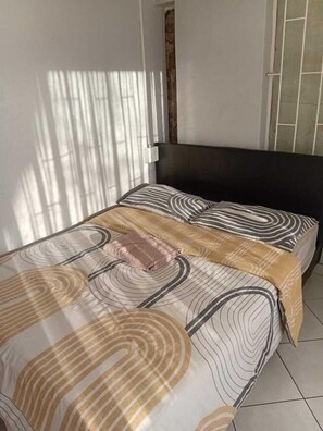 Una cama bien hecha con una colcha estampada y almohadas, un cabecero oscuro y una ventana con persianas.