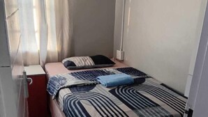 A small, well-lit bedroom with a single bed, a bedside table, and a window with sheer curtains.