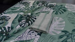 A bed with a tropical leaf pattern bedspread, a folded blanket, and a black chair in a room with a sheer curtain.