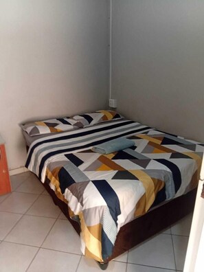 Una cama pequeña y bien hecha con una colcha de estampado geométrico en una habitación de paredes lisas y suelo de baldosas.