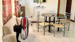 A living room with a sofa, a dining table with chairs, a TV mounted on the wall, and a standing fan. - Cranberry's 2Br Unit nr Convention Center #1 (Iloilo City)