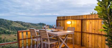 An outdoor wooden deck with a table and chairs, surrounded by a wooden fence, overlooking a hilly landscape.