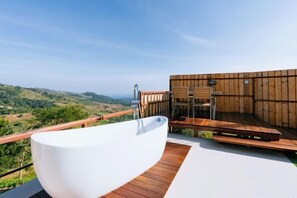 Une baignoire blanche, autoportante, posée sur une terrasse en bois, offrant une vue sur un paysage vallonné.