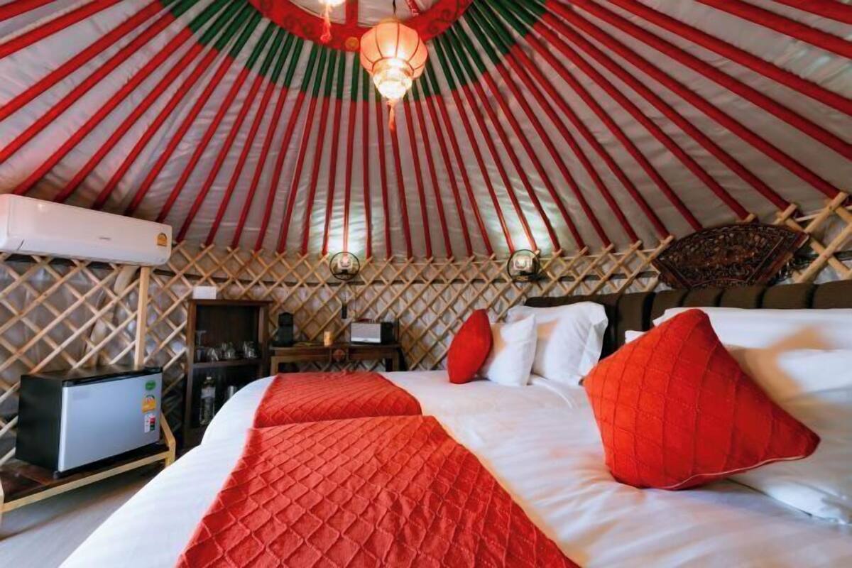 A room with a bed, red and white bedding, a red pillow, a small table with a lamp, and a wall-mounted air conditioner.