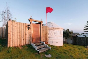 A wooden structure with a door and steps, a flag, and a white tent-like building in a grassy area.