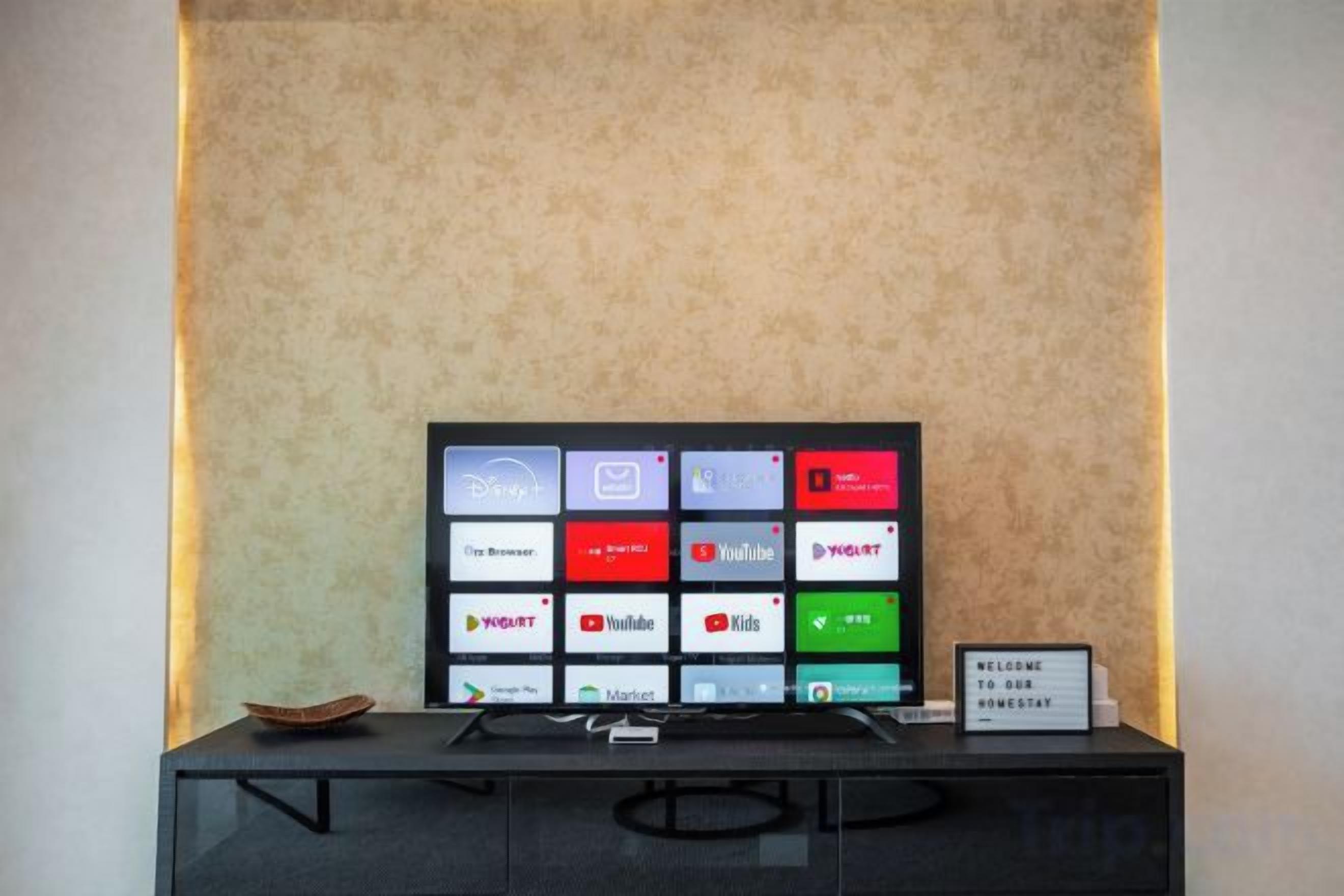 En platt-TV med en rutnätslayout av appikoner, placerad på ett svart stativ framför en vägg med en tydlig struktur.