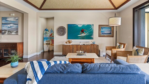 Een woonkamer met een grote bank, een bijzettafeltje en een televisie. Er staan surfplanken aan de muur en er hangt een schilderij met een strandthema.