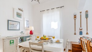 A dining area with a table set for two, a white cabinet with a first aid sign, and a window with sheer curtains. - #341 Casa da Guida | Central & Cozy (Albufeira)