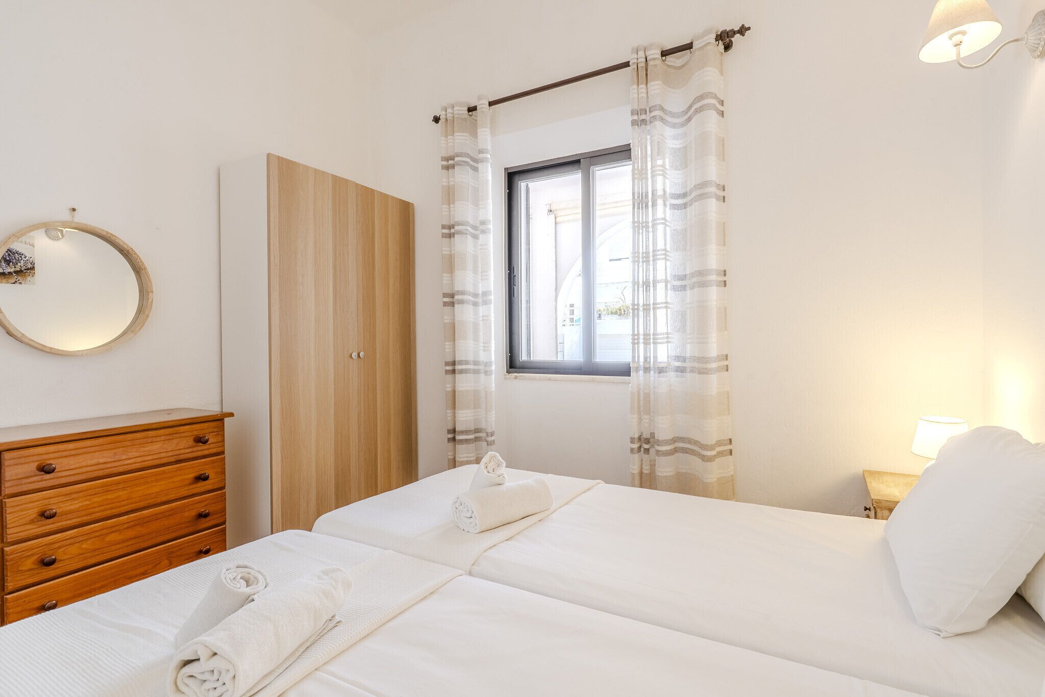 A bedroom with a bed, a wooden dresser, a mirror, and a window with curtains.