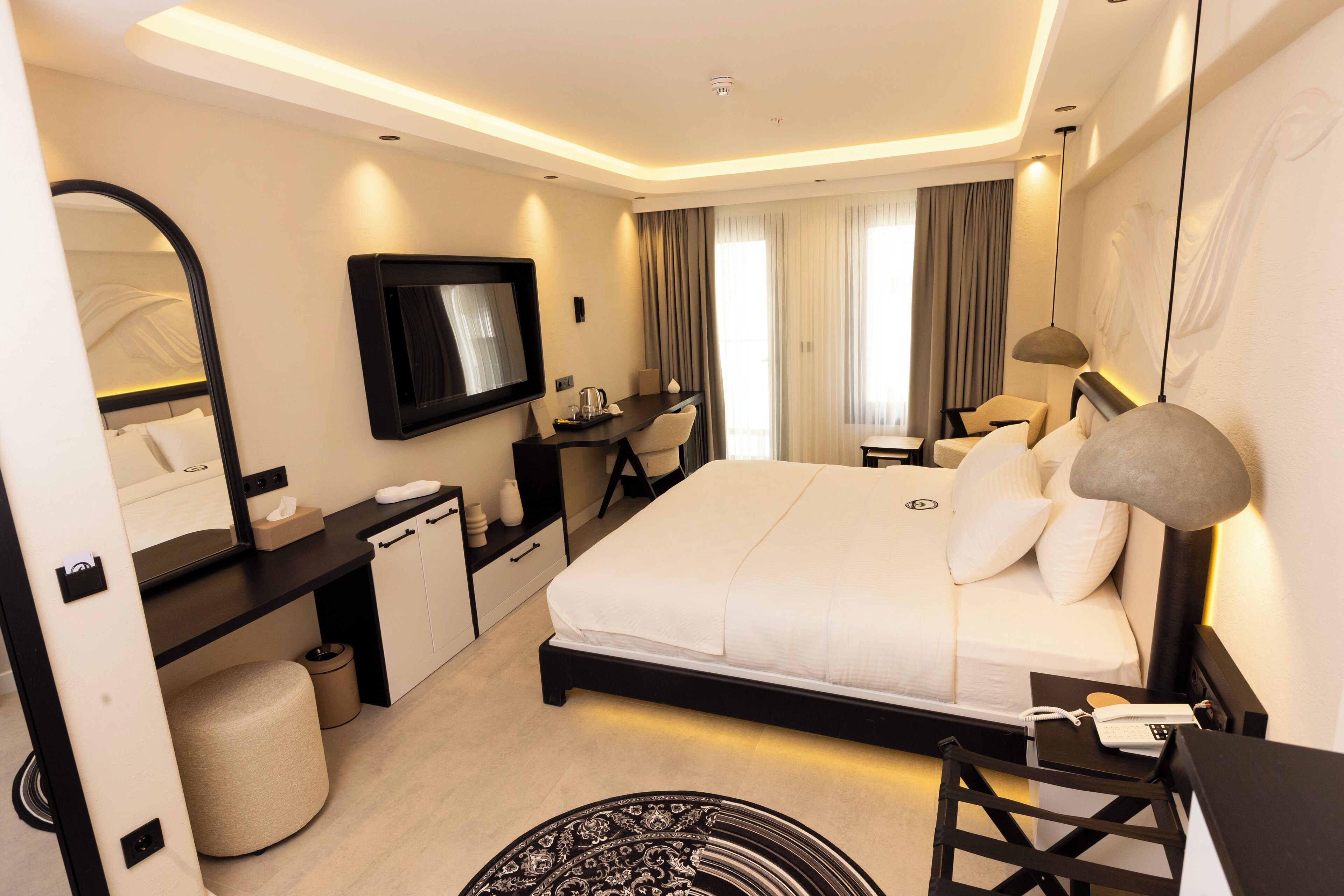 A modern hotel room with a large bed, a desk, a chair, a TV, and a lamp.
