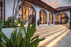 Une maison moderne avec un escalier en pierre menant à une entrée en porte de verre, le tout entouré de verdure.